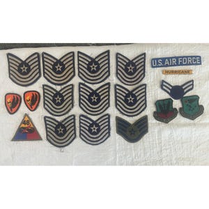 May include: A collection of vintage U.S. Air Force patches and insignia. Includes various rank chevrons with stars, wing emblems, and patches with text such as "U.S. AIR FORCE" and "HURRICANE". The patches are in shades of blue, gold, and red.