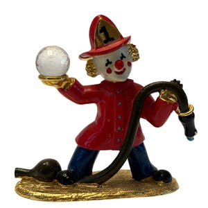 May include: A whimsical figurine of a clown dressed as a firefighter. The clown wears a red coat and hat with the number 1, blue pants, and holds a clear globe. A black hose and gold base complete the design.