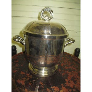 May include: A silver-colored metal ice bucket with a lid and two handles. The lid has a decorative handle. The bucket sits on a round base. The bucket is on a dark, patterned surface.