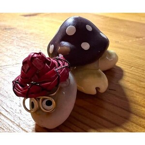 May include: A whimsical ceramic mushroom figurine with a dark brown cap and white spots. Beside it is a smaller, egg-shaped figure wearing a red woven hat and wire-rimmed glasses. The figures are on a wooden surface.