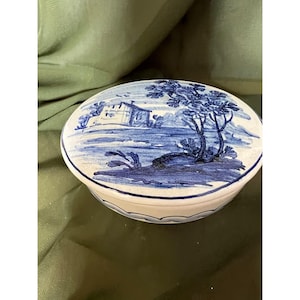 May include: Oval ceramic trinket box with a blue and white landscape design. The lid features a detailed scene of a building, trees, and water. The box has a scalloped edge and a white base.