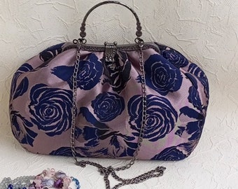 Może przedstawiać: Torebka w stylu vintage z kwiatowym wzorem. Torebka wykonana jest z miękkiej, fakturowanej tkaniny z ciemnoniebieskim wzorem kwiatowym na jasnoróżowym tle. Torebka ma metalową ramę i łańcuszkowy pasek.