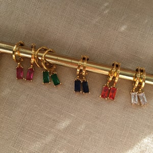 May include: A set of six gold hoop earrings with dangling rectangular gemstones. The gemstones are red, green, blue, red, clear and white.