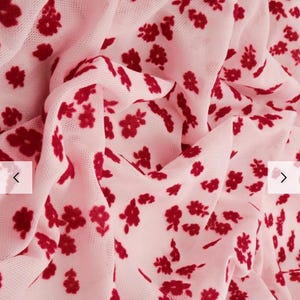 May include: A close-up of a sheer, pink fabric with a pattern of small, dark red flowers. The fabric is draped, creating soft folds and shadows. The floral design is scattered across the material.
