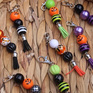 May include: A collection of Halloween-themed keychains featuring assorted beads in orange, black, purple, green, and white, including pumpkin-printed spheres, striped patterns, and glitter finishes, each finished with a silver lobster clasp and a tassel.