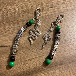 Op de afbeelding: Twee zilveren sleutelhangers met zwarte en groene kralen die "Slytherin" en "Malfoy" spellen. Elke sleutelhanger heeft een zilveren slangenbedel.