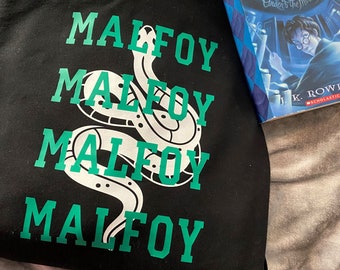 May include: Black hooded sweatshirt with the word "MALFOY" repeated four times in green text. A green snake with white scales is wrapped around the word "MALFOY". The sweatshirt is lying on a light gray surface. A copy of the book "Harry Potter and the Order of the Phoenix" is visible in the background.