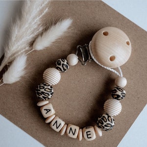 May include: A wooden baby teether with a clip and beads. The beads are brown and white with a leopard print pattern. The beads spell out the name "ANNE".