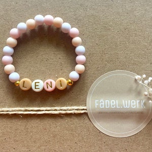 Könnte beinhalten: Ein rosa, weißes und beiges Perlenarmband mit dem Namen "LENI" in goldenen Buchstaben. Das Armband liegt auf einem braunen Hintergrund mit einem weißen runden Aufkleber, auf dem "Fädel.Werk LIEBEVOLL HANDGEMACHT |  EPZIGARTIG" steht.