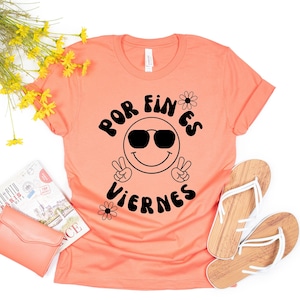 May include: A peach coloured t-shirt with a black graphic design that says "Por fin es viernes" with a smiley face wearing sunglasses and peace signs.