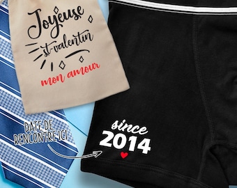 May include: A black boxer brief with a white waistband and the text "since 2014" printed on the leg. A small red heart is printed next to the year. A beige drawstring bag with the text "Joyeuse st-valentin mon amour" is in the background.