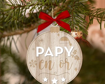 May include: Wooden Christmas ornament with the text "PAPY en or" in white and light brown. The ornament is round with a red bow and is hanging on a Christmas tree. The ornament has a snowflake, a hat, and three stars.