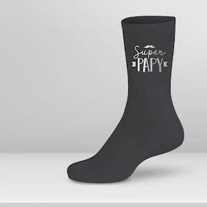Peut inclure: Chaussettes noires avec un motif argenté qui dit "Super Papy" avec une petite moustache au-dessus du texte.