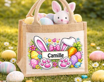 May include: A beige burlap Easter tote bag with a bunny graphic and the name "Camille". The bag is filled with colorful Easter eggs and a plush bunny. The bag is sitting on green grass with scattered Easter eggs.