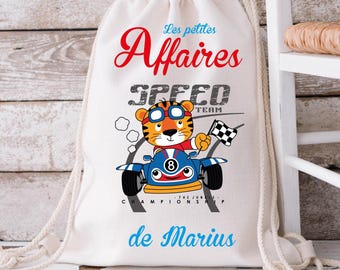 May include: A white drawstring bag with the text "Les petites Affaires" in red script above "SPEED TEAM" in gray block letters. A cartoon tiger in goggles drives a blue race car, holding a checkered flag. The text "de Marius" is below.