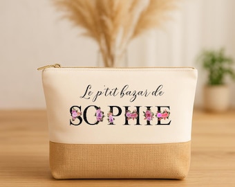 May include: A beige and tan cosmetic bag with a gold zipper. The text "Le p'tit bazar de SOPHIE" is printed on the front, with the name "SOPHIE" in large black letters decorated with pink and purple flowers. The bag is rectangular.