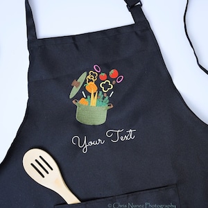 May include: Black kitchen apron with a pocket and a design of a pot with vegetables and the text "Your Text" embroidered on it.