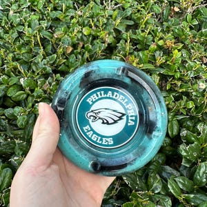 May include: Round ashtray with a teal and black design. The center features the Philadelphia Eagles logo in white and teal. The ashtray is held against a backdrop of green foliage.