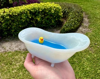 May include: A small, white bathtub filled with blue liquid and a yellow rubber ducky. The miniature tub has small white feet and is held in a person's hand. The background features green grass, bushes, and a sidewalk.