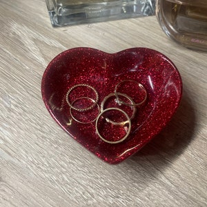 May include: A heart-shaped, red glitter resin trinket dish holding several gold rings. The dish is on a light wood surface, with two perfume bottles in the background.