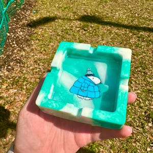 May include: Square ashtray with a teal and white marbled design. The ashtray features a cartoon character in a blue coat and hat. The ashtray is held in a hand.
