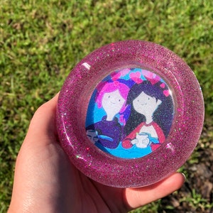 May include: A round, glittery pink resin ashtray. The ashtray features a cartoon image of two characters holding mugs. The background of the image is blue. The ashtray is held in a hand against a green background.