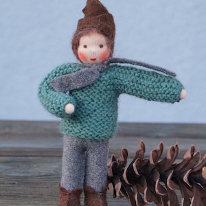 Könnte beinhalten: Handgemachte Puppe mit einem gestrickten tealfarbenen Pullover, grauer Hose und einem braunen Hut und Stiefeln. Die Puppe hat einen grauen Schal und ein bemaltes Gesicht mit rosigen Wangen. Ein großer Tannenzapfen liegt in der Nähe der Puppe.