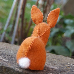 Könnte beinhalten: Ein kleiner orangefarbener Filzhase mit einem weißen Filzschwanz. Der Hase sitzt auf einem Stein.