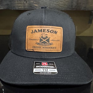 May include: Black trucker hat with a brown leather patch that says "Jameson Irish Whiskey" and a black and white logo. The hat also has a black tag that says "Richardson 112 Original Trucker Adjustable".