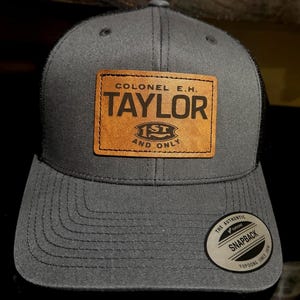 May include: Gray and black trucker hat with a brown leather patch that says "Colonel E.H. Taylor 1st and Only". The hat has a black mesh back and a black snap closure.