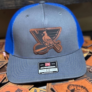 May include: A gray and blue trucker hat with a brown leather patch featuring a cardinal bird and the text "St. Louis Blues". The hat is adjustable and has the Richardson logo and the text "Original 112 Trucker" on the back.