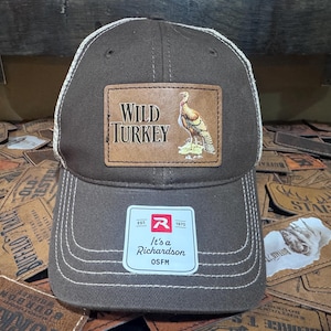 May include: Brown baseball cap with a white mesh back and a leather patch that reads "WILD TURKEY" with a turkey illustration. The cap has a curved brim and a white stitched design. The tag reads "It's a Richardson OSFM".