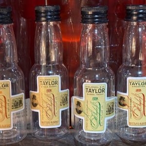 May include: Four clear glass bottles of Colonel E.H. Taylor small batch bourbon whiskey. The bottles are labelled with the brand name and the type of whiskey. The bottles are all in a row and are all the same size.