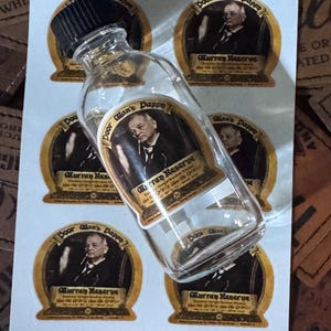 May include: A clear glass bottle with a black cap, resting on a sheet of stickers. The stickers feature a vintage design with the text "Door Man's Dappe" and an image of a man. The stickers are brown and gold.