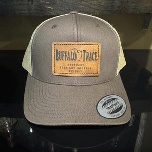 May include: A brown and tan trucker hat with a brown leather patch that says "Buffalo Trace Kentucky Straight Bourbon Whiskey". The hat has a black snap closure and a white "The Authentic Snapback" label.