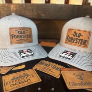 May include: Two gray and black trucker hats with brown leather patches that say "Old Forester" on them. The hats are sitting on a table with other leather patches.