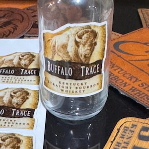 May include: A clear glass bottle with a label that says "Buffalo Trace Kentucky Straight Bourbon Whiskey". The label features a brown and white illustration of a buffalo.