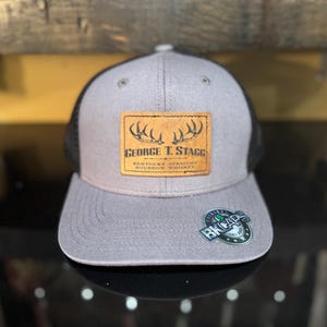 Puede incluir: Una gorra de camionero gris y negra con un parche de cuero marrón que presenta una cornamenta de ciervo y el texto "George T. Stagg Kentucky Straight Bourbon Whiskey". La gorra también tiene una pegatina en blanco y negro con el texto "BK Caps" en la parte delantera.