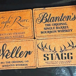 May include: Four brown leather coasters with black text. The coasters feature the names of different bourbon whiskeys: Eagle Rare, Blanton's, Weller, and Stagg.