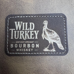May include: A rectangular, dark gray leather patch with white stitching around the edges. The patch features the words "WILD TURKEY" above "BOURBON WHISKEY" and a detailed illustration of a turkey.