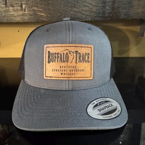 May include: A gray and black trucker hat with a brown leather patch that says "Buffalo Trace Kentucky Straight Bourbon Whiskey". The hat has a black mesh back and a snap closure.