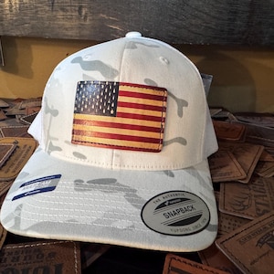 May include: White camouflage baseball cap with a leather American flag patch. The cap features a curved brim and a snapback closure. The flag patch has a distressed look, with red and brown stripes and a blue field of stars.