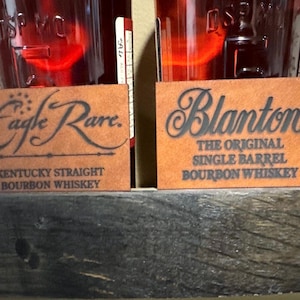 May include: Two brown leather labels with black text. The top label reads "Eagle Rare. Kentucky Straight Bourbon Whiskey". The bottom label reads "Blanton's The Original Single Barrel Bourbon Whiskey".