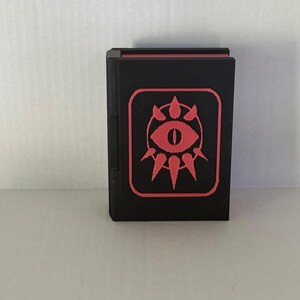 May include: A black book with a red eye symbol on the cover. The symbol is surrounded by a circle of spikes.