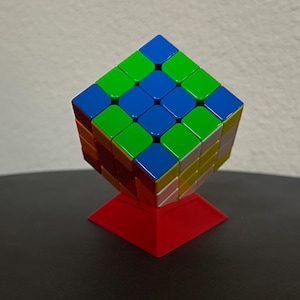 May include: A multi-colored cube puzzle with green, blue, red, yellow, and orange squares. The cube is displayed on a red stand, set against a neutral background. The cube is a classic brain teaser.