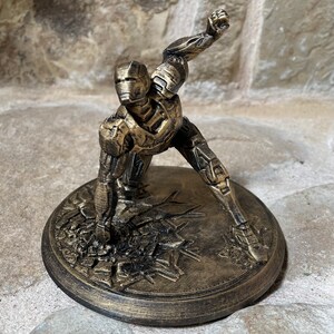 May include: A gold-coloured 3D printed figurine of a superhero in a fighting pose. The figurine is standing on a round base with a textured surface.