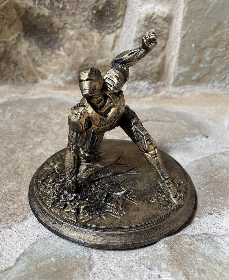 May include: A gold-coloured 3D printed figurine of a superhero in a fighting pose. The figurine is standing on a round base with a textured surface.