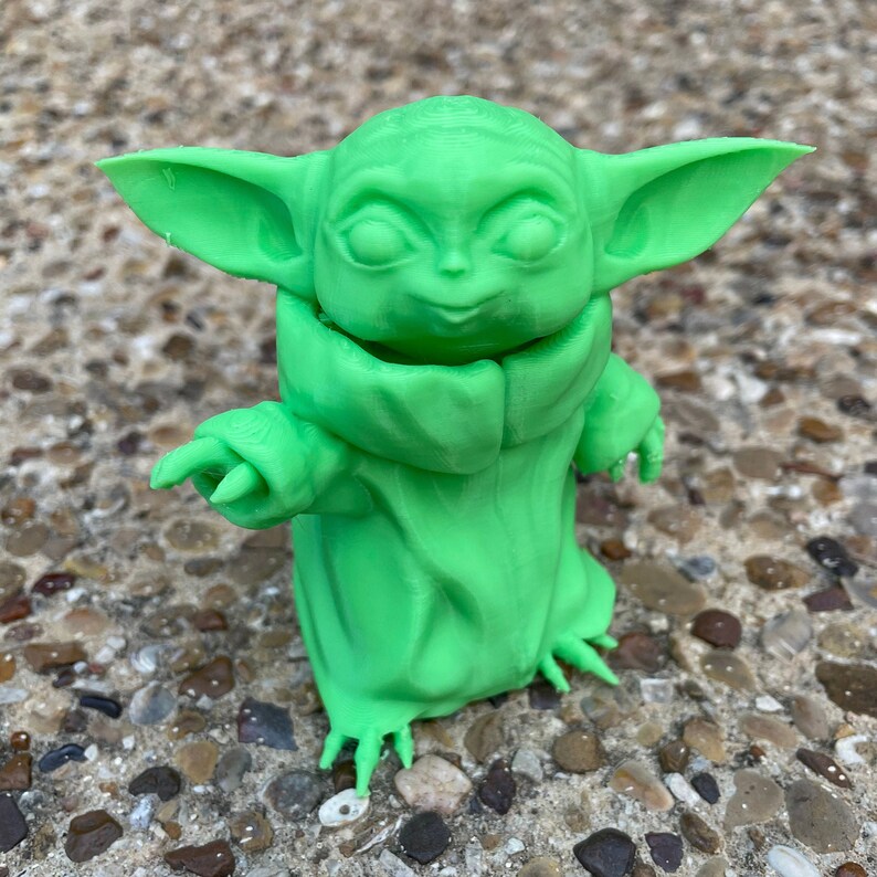 May include: A bright green figurine of a character with large ears and a hooded cloak. The figure has a small, pointed hand extended. The figurine is made of a solid material and is standing on a surface of small rocks.