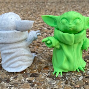 May include: Two 3D-printed figurines of a character with large ears and a robe. One is speckled gray, the other is bright green. Both are posed with one arm extended, set against a natural, textured background.