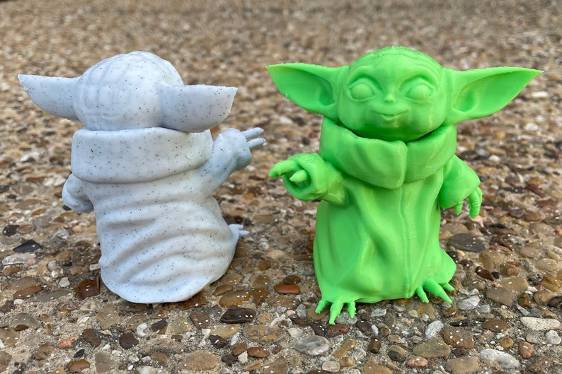 May include: Two 3D-printed figurines of a character with large ears and a robe. One is speckled gray, the other is bright green. Both are posed with one arm extended, set against a natural, textured background.